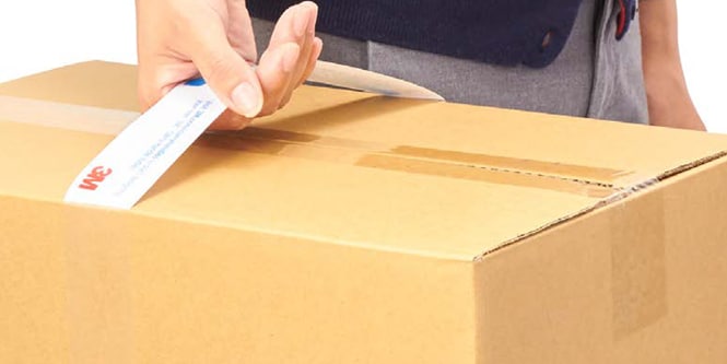 Close-up of a person applying a white 3M‑branded strip across the top seam of a sealed brown cardboard box.
