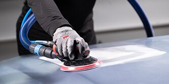 Worker wearing protective gloves operating a 3M orbital sander with a red-backed disc to smooth a vehicle surface.
