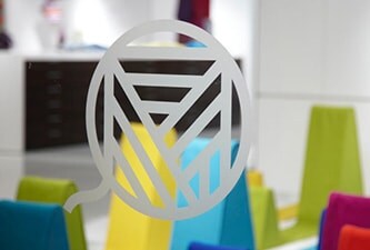 Fenêtre en verre limpide ornée d’un logo gravé en relief, sublimé par des formes colorées en arrière-plan.
