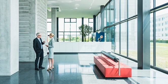 Vue panoramique du hall d’entrée d’un immeuble de bureaux contemporain, bordé de parois vitrées où deux hommes d’affaires échangent.
