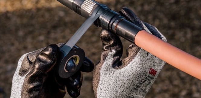 Deux mains gantées enroulent du ruban électrique noir autour d’un câble.
