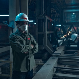 A worker stands in an industrial setting, wearing a white hard hat with the 3M logo and a face mask.