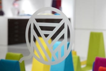 Fenêtre en verre limpide ornée d’un logo gravé en relief, sublimé par des formes colorées en arrière-plan.
