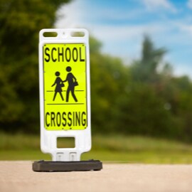 Standalone sign with yellow background and black image of two people crossing street and words 'school crossing'.