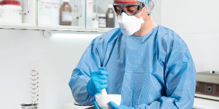 A person in blue scrubs, a surgical mask, protective goggles, and a cap is standing in a laboratory or medical environment.
