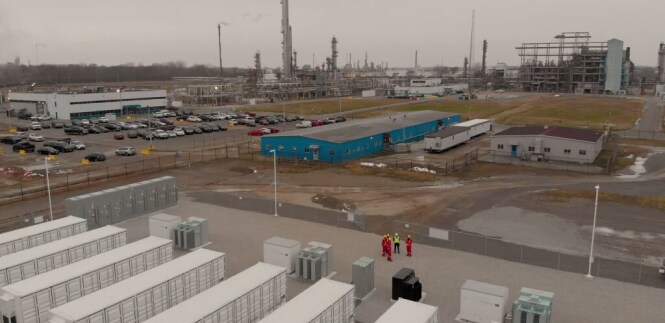 An industrial complex with several large buildings and structures inluiding a parking lot filled with cars.
