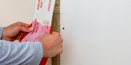 A person applying a red and white firestop tape to a wall seam, ensuring proper sealing and fire protection.
