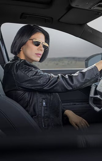Un individu vêtu d'une veste en cuir et portant des lunettes de soleil est assis au volant d'une voiture, prêt à démarrer. 
