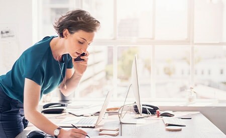Un individu au téléphone, debout au-dessus d’un bureau, prend des notes dans un espace blanc inondé de lumière, agrémenté de vastes fenêtres.
