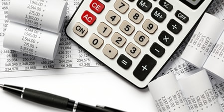 Grande calculatrice posée sur des feuilles de calcul financières, entourée de reçus en rouleau et d’un stylo, suggérant le suivi comptable et des dépenses.
