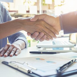 Gros plan sur deux professionnels se serrant la main à un bureau, entourés de graphiques, de stylos et d’un ordinateur portable ouvert, illustrant la conclusion d’un accord commercial.