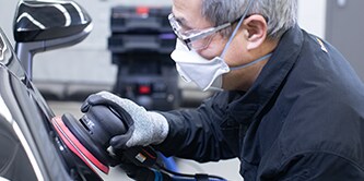 Worker wearing safety glasses, gloves, and a protective mask using a handheld power buffer to polish the surface of a vehicle.
