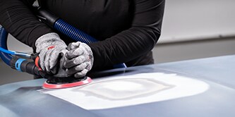 Un ouvrier portant des gants de protection utilise une ponceuse orbitale équipée d’un disque à dos rouge pour lisser une zone préparée de la surface d’un véhicule.
