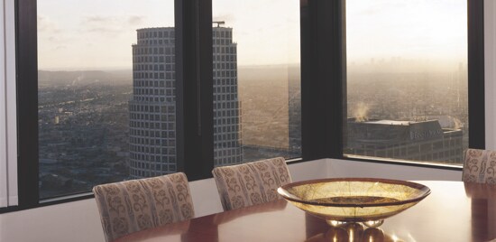 Une table de salle à manger avec une ville au crépuscule visible à travers des grandes fenêtres.
