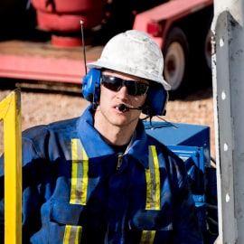 A person wearing a white hard hat and blue protective coveralls with a yellow reflective stripes is equipped with blue earmuffs.
