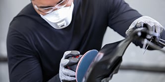 Un ouvrier équipé de gants de protection utilise une ponceuse électrique munie d’un disque abrasif bleu sur la surface d’un véhicule.
