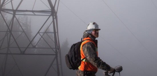 Un travailleur portant un casque de protection et un gilet réfléchissant avec des lignes électriques en arrière-plan.
