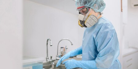 A person in medical protective gear, including a blue gown, gloves, a surgical mask, and a face shield, stands in a clinical setting with a sink in the background.

