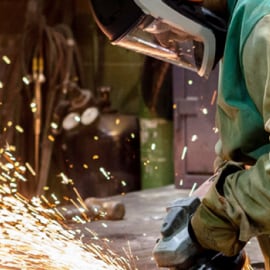 A worker wearing protective gear, including a face shield and gloves is operating a grinding tool.