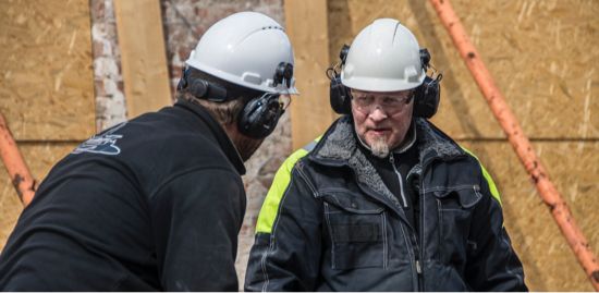 Two construction workers wearing hearing protection, talking to each other.
