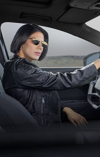 An individual in  leather jacket and sunglasses sits in the driver's seat of a car, ready to drive. 
