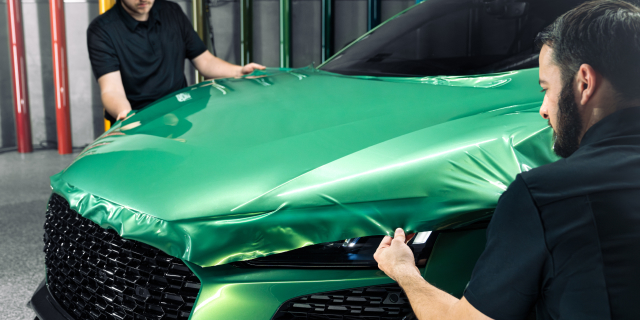 Deux installateurs recouvrent le capot d'une voiture sport d'une pellicule d’habillage de couleur vert émeraude.

