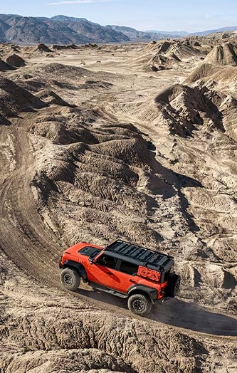 VUS recouvert d’une pellicule d'habillage rouge roulant sur une piste tout-terrain accidentée dans le désert, avec des montagnes à l'horizon.
