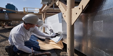 Workers wearing hard hats applying protective wrap to industrial equipment outdoors, with metal structures and machinery in the background.
