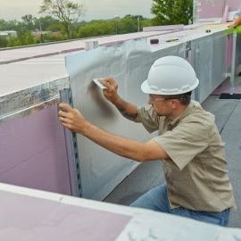 A construction worker wearing a hard hat is applying a sheet of insulation material to a building structure.
