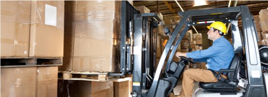A warehouse worker wearing a hard hat, driving a fork lift that is lifting up a pallet of boxes.
