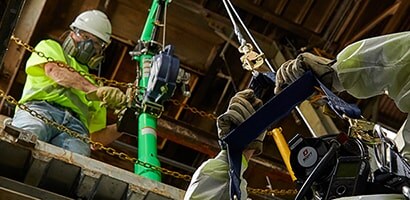 A construction worker wearing a high-visibility vest and safety helmet is using a green vertical jackhammer to break up concrete.
