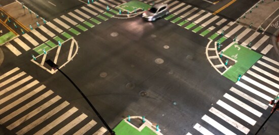 Overhead view of car driving through intersection with crosswalk lines and green bike lane lines.
