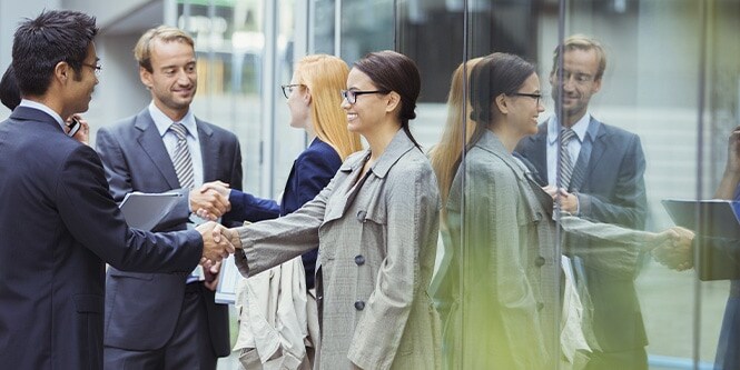 Un groupe d’hommes d’affaires échange une poignée de main devant une large baie vitrée d’un immeuble de bureaux.
