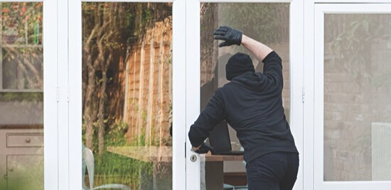 Un cambrioleur vêtu tout en noir qui porte des gants et un masque et qui regarde à travers d'une porte vitrée.
