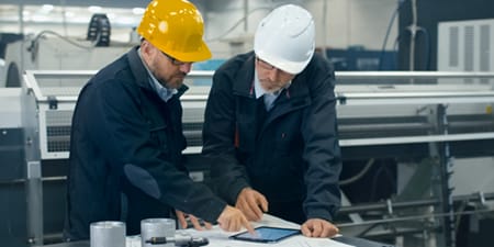 Deux ouvriers industriels portant des casques jaunes et blancs examinent des plans et une tablette à un poste de travail, tandis que les machines d’usine se trouvent en arrière-plan.
