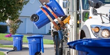 Automated side‑loader recycling truck emptying blue curbside bins on a suburban street, mechanical arm tipping one container while others line the curb.
