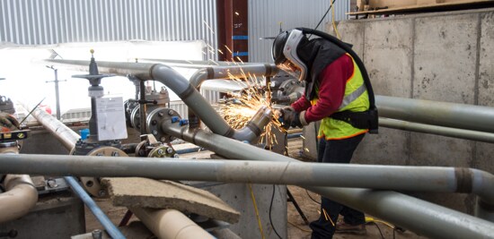 A metalworker wearing a safety vest and PAPR, grinding a metal pipe outside.
