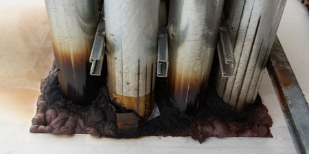 Tuyaux en métal avec rouille et corrosion visibles à la base, entourés d’une substance sombre et mousseuse sur une surface plane.
