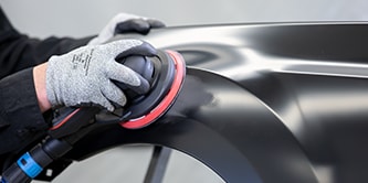 Worker wearing protective gloves using an orbital sander with a red-backed disc to smooth a black vehicle panel.
