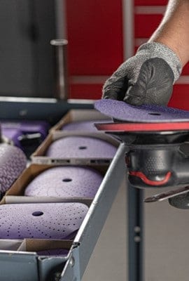 Une personne portant un gant noir tient un disque abrasif perforé violet et l’attache à une ponceuse orbitale avec des abrasifs organisés en arrière-plan.