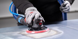 Un ouvrier portant des gants de protection utilise une ponceuse orbitale 3M équipée d’un disque à dos rouge pour lisser la surface d’un véhicule.
