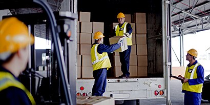 Workers in hard hats and yellow safety vests unloading boxes from a delivery truck.
