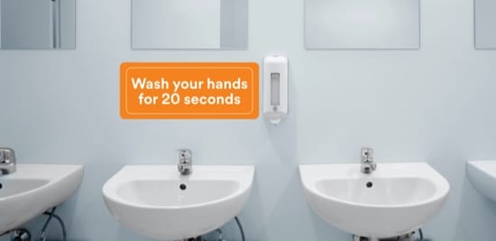 Une salle de bain publique avec lavabos et miroirs, et une pancarte recommandant de se laver les mains pendant 20 secondes.
