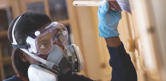 A woman painting indoors wearing a respirator and safety goggles.
