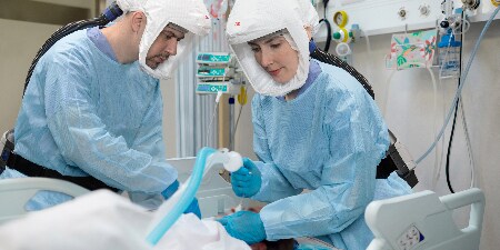 Two healthcare professionals in blue scrubs and protective gear, including gloves and face shields, are attending to a patient in a clinical setting.
