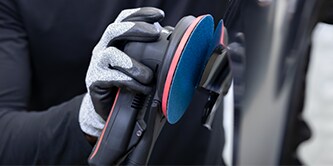 Ouvrier portant des gants de protection tenant une ponceuse électrique équipée d’un disque abrasif bleu, prêt à intervenir sur la surface d’un véhicule.
