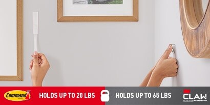 Two hands are shown installing wall-hanging hardware, the left hand is holding a Command strip, while the right hand is using a 3M CLAW drywall picture hanger.
