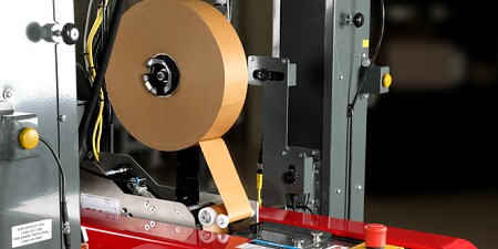 Machine industrielle pour sceller des cartons, équipée d’un grand rouleau de ruban en papier brun passant dans des rouleaux, posée sur une base rouge avec boutons de commande et structure métallique.
