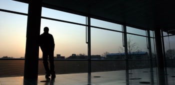 Un homme appuyé contre un pilier regarde le soleil se coucher par des baies vitrées.
