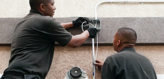 Deux électriciens acheminent des fils sur le côté d’un bâtiment.
